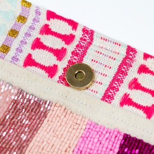 May include: A close-up of a pink and white beaded bag with a gold metal clasp. The bag has a woven fabric flap with a pink and white geometric pattern.