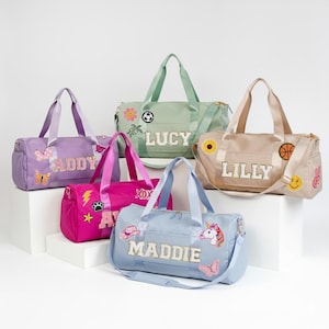 May include: Five duffel bags in lavender, sage green, beige, hot pink, and light blue. Each bag features a personalized name in white glittery letters and various colorful embroidered patches including butterflies, sports, and animals.