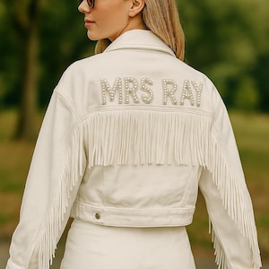 May include: An off-white denim jacket featuring long fringe detailing on the back and sleeves, with the text MRS RAY spelled out in white pearl-like beads across the upper back, paired with matching off-white denim shorts.