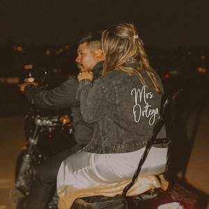 Könnte beinhalten: Ein Paar auf einem Motorrad bei Nacht. Die Frau trägt eine dunkelgraue Jeansjacke mit "Mrs. Ortega" in weißer Schrift auf dem Rücken. Sie trägt eine weiße Hose und einen beigen Rock. Der Mann trägt eine schwarze Jacke.