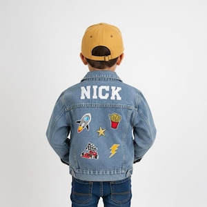 Può includere: Una giacca di jeans azzurra con il nome NICK ricamato in bianco sulla parte superiore della schiena. La giacca è decorata con toppe colorate che raffigurano un razzo, una porzione di patatine fritte, una stella gialla, un'auto e un fulmine.