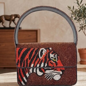 May include: Brown beaded handbag with a black handle and a flap closure. The front features a tiger design in red, black, white, and yellow beads. The bag is sitting on a marble table.