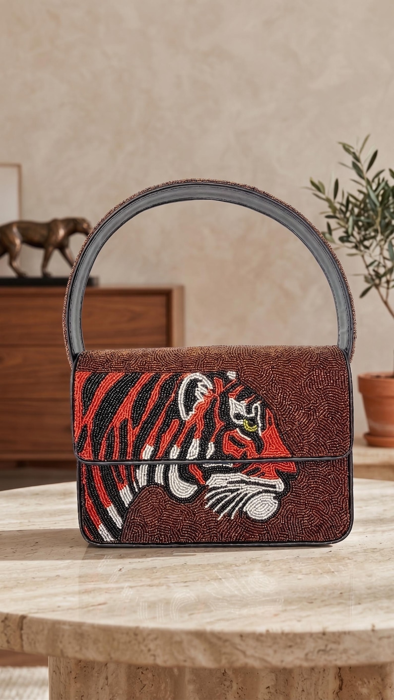 May include: Brown beaded handbag with a black handle and a flap closure. The front features a tiger design in red, black, white, and yellow beads. The bag is sitting on a marble table.