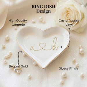 May include: A heart-shaped white ceramic ring dish featuring a glossy finish and an elegant gold trim. The center displays gold script lettering with the letters a and k connected by a heart symbol. Text includes RING DISH Design, High Quality Ceramic, Customizable Vinyl, Elegant Gold Trim, and Glossy Finish.