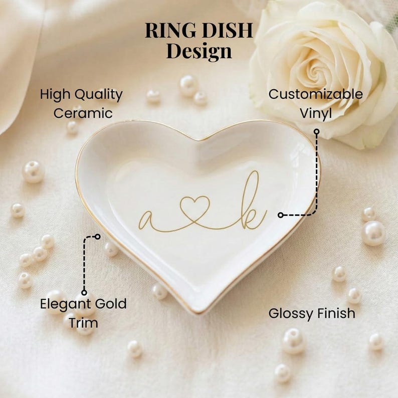 May include: A heart-shaped white ceramic ring dish featuring a glossy finish and an elegant gold trim. The center displays gold script lettering with the letters a and k connected by a heart symbol. Text includes RING DISH Design, High Quality Ceramic, Customizable Vinyl, Elegant Gold Trim, and Glossy Finish.