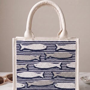 May include: A white canvas tote bag with a beaded design of fish swimming across navy blue and white striped fabric. The bag has a rectangular shape with a sturdy handle and a neutral color palette.