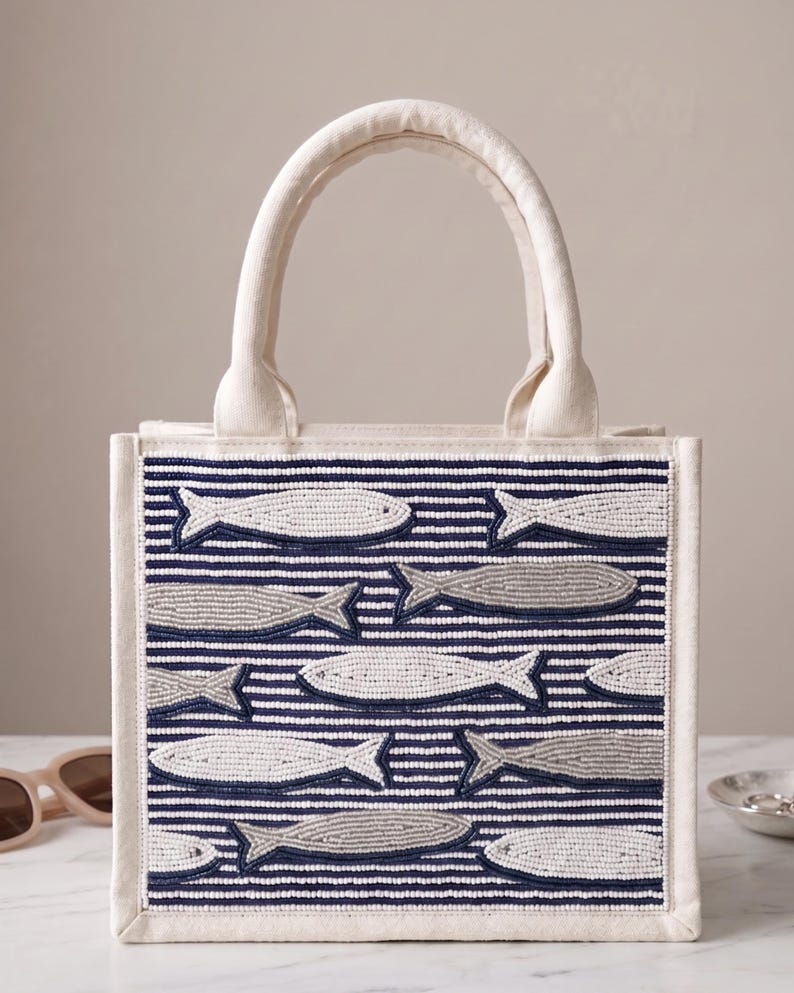 May include: A white canvas tote bag with a beaded design of fish swimming across navy blue and white striped fabric. The bag has a rectangular shape with a sturdy handle and a neutral color palette.