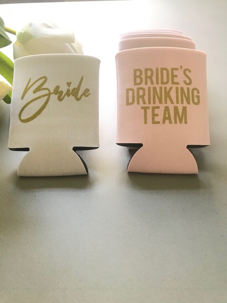 May include: Two white foam beverage insulators. One is white with gold lettering that says "Bride". The other is pink with gold lettering that says "Bride's Drinking Team".