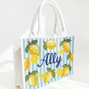 May include: White tote bag with blue and white striped pattern. The bag features embroidered yellow lemons and the name "Ally" in blue script. The bag has white handles and a rectangular shape.