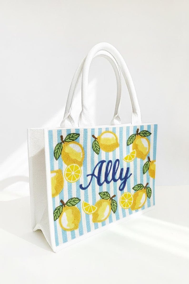 May include: White tote bag with blue and white striped pattern. The bag features embroidered yellow lemons and the name "Ally" in blue script. The bag has white handles and a rectangular shape.