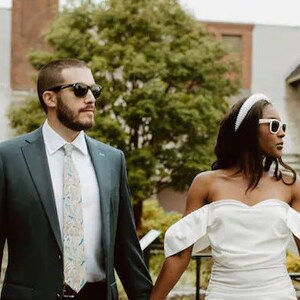 May include: A couple in formal attire, the man wearing a dark blue suit with a patterned tie and sunglasses, the woman wearing a white off-the-shoulder dress with a white headband and sunglasses.