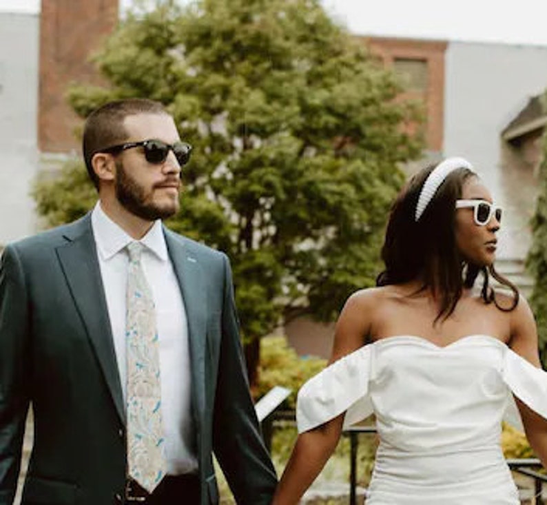 May include: A couple in formal attire, the man wearing a dark blue suit with a patterned tie and sunglasses, the woman wearing a white off-the-shoulder dress with a white headband and sunglasses.