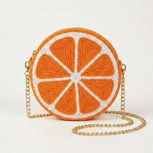 May include: A round crossbody bag shaped like a sliced orange, featuring vibrant orange and white glass beads arranged in a citrus segment pattern. The bag includes a gold-tone metal chain strap attached to the sides with lobster claw clasps.