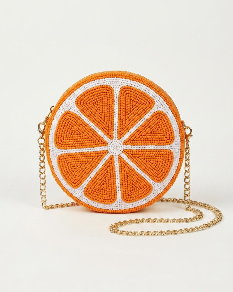 May include: A round crossbody bag shaped like a sliced orange, featuring vibrant orange and white glass beads arranged in a citrus segment pattern. The bag includes a gold-tone metal chain strap attached to the sides with lobster claw clasps.