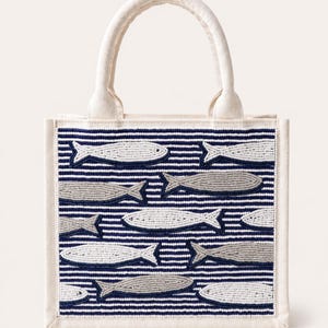 May include: A small, off-white tote bag with a white handle. The bag features a navy blue and white striped pattern with beaded fish in shades of gray and white. The bag has a rectangular shape.