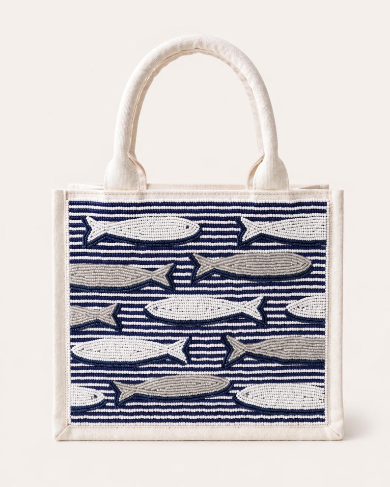 May include: A small, off-white tote bag with a white handle. The bag features a navy blue and white striped pattern with beaded fish in shades of gray and white. The bag has a rectangular shape.