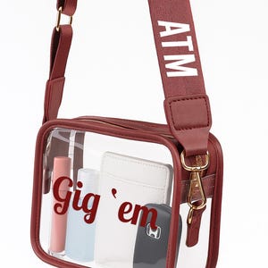 A maroon crossbody bag with a clear front pocket, displaying various items such as a wallet, keys, and a phone.