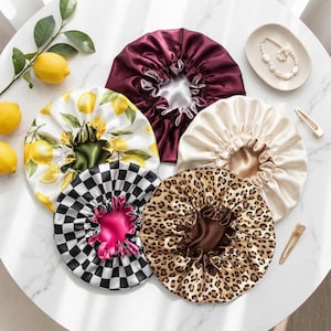 May include: A collection of five circular satin hair bonnets featuring diverse patterns including a lemon print, a black and white checkerboard, a leopard print, a solid burgundy, and a solid cream color, each with a gathered elastic edge.