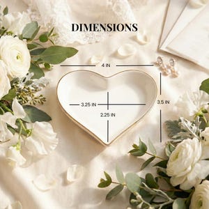 May include: A white heart-shaped ceramic trinket dish with a gold-toned rim. The dish features printed dimension lines indicating a width of 4 inches, a height of 3.5 inches, an inner width of 3.25 inches, and an inner depth of 2.25 inches.