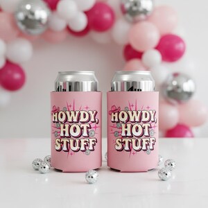 Puede incluir: Dos enfriadores de latas rosas con el texto "HOWDY HOT STUFF" en una fuente retro, adornados con un sombrero de vaquero y detalles brillantes. Los enfriadores están sobre una superficie blanca con decoraciones de bolas de discoteca.