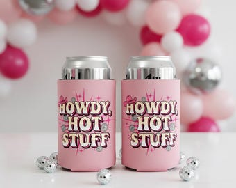 May include: Two pink can coolers with the text "HOWDY HOT STUFF" in a retro font, adorned with a cowboy hat and sparkling accents. The coolers are on a white surface with disco ball decorations and a balloon arch in the background.