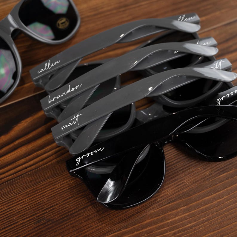 May include: Five pairs of sunglasses with personalized names on the frames. The sunglasses are gray and black. The names are "groom", "matt", "brandon", "callen", and "ellen".