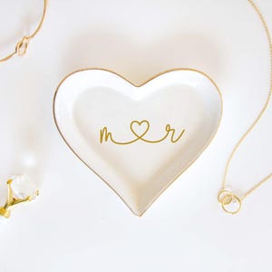 May include: A white heart-shaped ceramic trinket dish with a gold-colored rim. The center of the dish features a gold-colored graphic design consisting of the lowercase letters m and r connected by a heart shape in the middle.