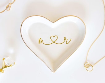 Può includere: Un piattino in ceramica a forma di cuore bianco con un bordo dorato. Il centro del piatto presenta un design grafico dorato composto dalle lettere minuscole m e r collegate da una forma a cuore nel mezzo.