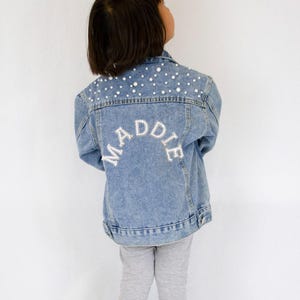 May include: Light blue denim jacket with pearl embellishments on the shoulders. The name "MADDIE" is embroidered in white across the back. A stylish and personalized children's jacket.