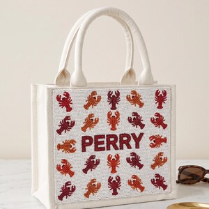 May include: Off-white canvas tote bag with two handles. The front of the bag is decorated with beaded red, orange, and maroon lobsters. The name "PERRY" is spelled out in maroon beads. A stylish and unique accessory.