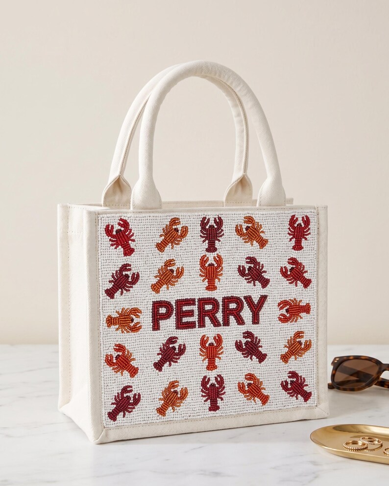 May include: Off-white canvas tote bag with two handles. The front of the bag is decorated with beaded red, orange, and maroon lobsters. The name "PERRY" is spelled out in maroon beads. A stylish and unique accessory.