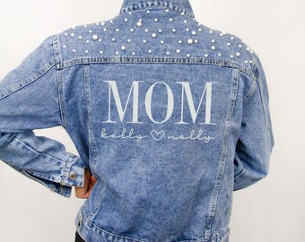 Könnte beinhalten: Eine hellblaue Jeansjacke mit weißen Perlenverzierungen am oberen Rücken und an den Schultern. Auf der Rückseite befindet sich der weiße Schriftzug MOM in einer großen Serifenschrift, gefolgt von dem kursiven Text kelly Herz molly.