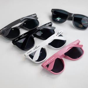 May include: Five pairs of sunglasses with personalized names on the arms. The sunglasses are in black, white, and pink frames. The names on the sunglasses are Jordan, Mr. Allen, Mrs. Allen, McKenzie, and Jessie.