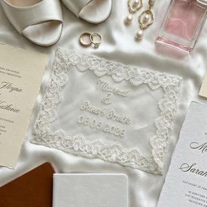 May include: A collection of wedding accessories including white satin heels, gold rings, pearl earrings, and a perfume bottle. A white lace invitation with embroidered names and date is centered. Brown and white vow books are also present.