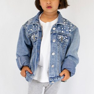 K&ouml;nnte beinhalten: Eine hellblaue Jeansjacke mit klassischem Kragen, langen &Auml;rmeln und silberfarbenen Metallkn&ouml;pfen. Die oberen Vorderteile und Brusttaschen sind mit zahlreichen wei&szlig;en Kunstperlen in verschiedenen Gr&ouml;&szlig;en verziert.