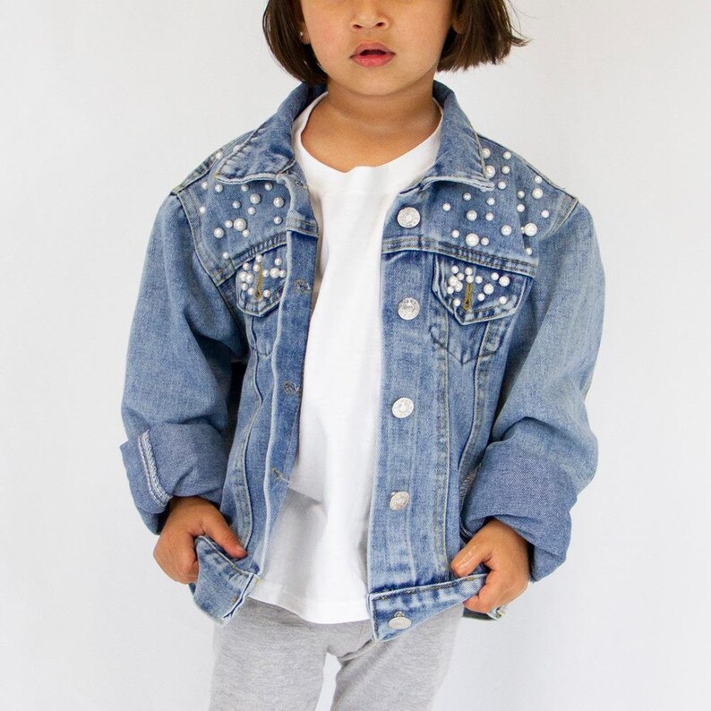 K&ouml;nnte beinhalten: Eine hellblaue Jeansjacke mit klassischem Kragen, langen &Auml;rmeln und silberfarbenen Metallkn&ouml;pfen. Die oberen Vorderteile und Brusttaschen sind mit zahlreichen wei&szlig;en Kunstperlen in verschiedenen Gr&ouml;&szlig;en verziert.