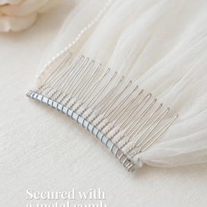 A silver metal comb rests on a white fabric, secured with a pearl bead.