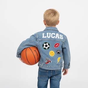 May include: A light blue denim jacket featuring the name LUCAS in white block letters across the back. The jacket is decorated with embroidered patches including a soccer ball, football, smiley face, paw print, race car, and yellow lightning bolt.