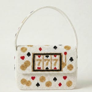 Könnte beinhalten: Eine weiße, perlenbesetzte Handtasche mit einer zentralen Spielautomaten-Grafik, die die Zahl 777 zeigt. Die Tasche ist mit goldenen Münzmotiven sowie roten und schwarzen Spielkartensymbolen wie Herz, Karo, Pik und Kreuz verziert.