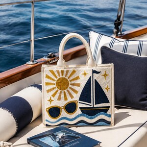 May include: A square-shaped cream-colored tote bag featuring intricate beadwork. The design displays a golden sun, a dark blue sailboat, light blue wavy lines representing water, and a pair of sunglasses, all accented with small golden star shapes.