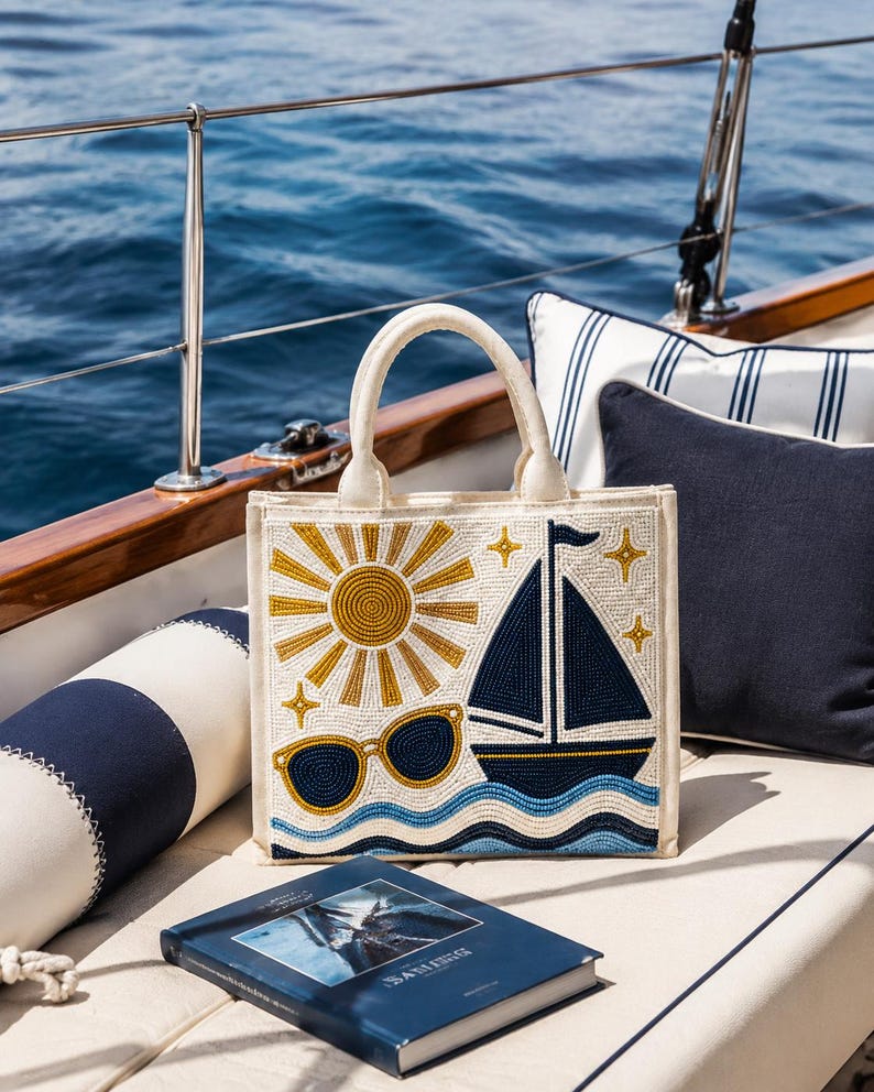 May include: A square-shaped cream-colored tote bag featuring intricate beadwork. The design displays a golden sun, a dark blue sailboat, light blue wavy lines representing water, and a pair of sunglasses, all accented with small golden star shapes.