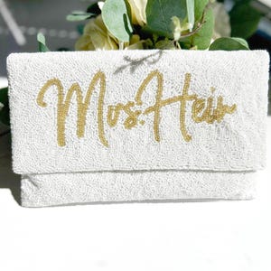 May include: A rectangular white beaded clutch bag featuring the gold beaded text "Mrs. Heir" in a cursive script font. The exterior is covered in small white beads, and the lettering is composed of metallic gold beads across the front flap.