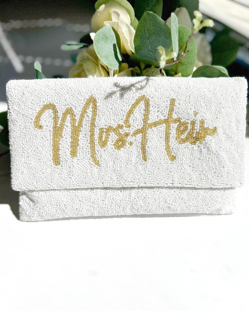 May include: A rectangular white beaded clutch bag featuring the gold beaded text "Mrs. Heir" in a cursive script font. The exterior is covered in small white beads, and the lettering is composed of metallic gold beads across the front flap.