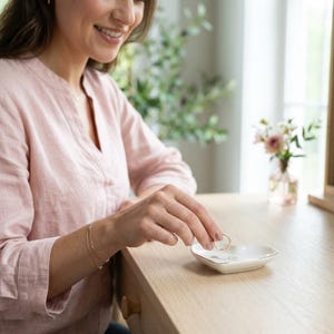 May include: A small, white, heart-shaped ceramic trinket dish with a gold-colored rim. The dish features a delicate, thin black line drawing of a heart and a script letter on its surface, designed to hold small jewelry items like rings.