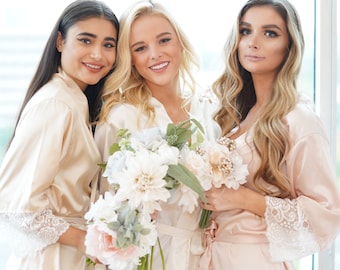 May include: Three women in satin robes, in champagne and blush tones, with lace detailing on the sleeves. Each holds a bouquet of white, pink, and blue flowers. The lighting is soft and natural. The image suggests a bridal or celebratory theme.