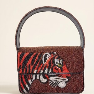 May include: A beaded handbag with a tiger design. The bag features a brown background with a tiger face in red, black, and white beads. The handle is a dark brown color. The bag has a rectangular shape.