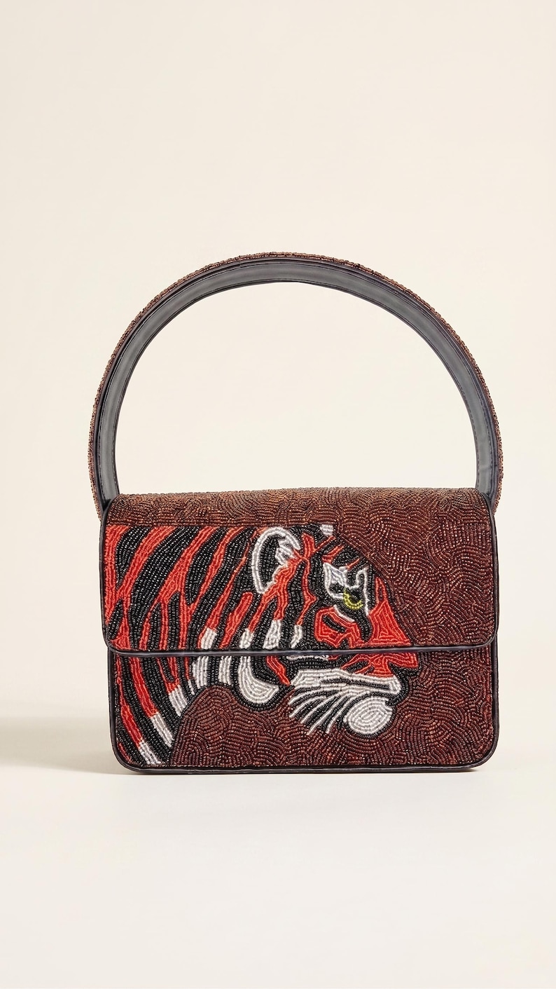 May include: A beaded handbag with a tiger design. The bag features a brown background with a tiger face in red, black, and white beads. The handle is a dark brown color. The bag has a rectangular shape.