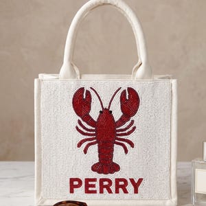 May include: A white beaded tote bag with a red lobster design and the name "PERRY" in red letters. The bag has a cream-colored handle and trim.