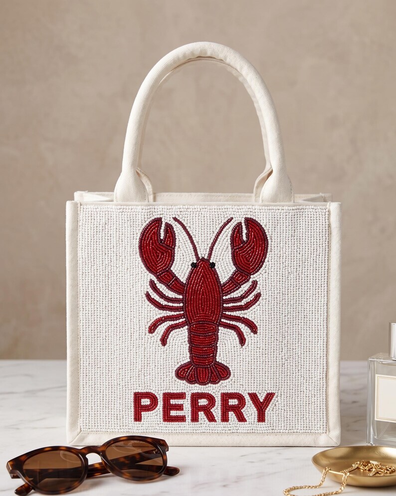 May include: A white beaded tote bag with a red lobster design and the name "PERRY" in red letters. The bag has a cream-colored handle and trim.