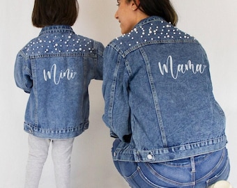 Könnte beinhalten: Zwei blaue Jeansjacken mit weißen Perlenverzierungen am oberen Rücken und an den Schultern. Die kleinere Jacke trägt den Schriftzug Mini in weißer Schreibschrift, die größere Jacke den Schriftzug Mama in weißer Schreibschrift.
